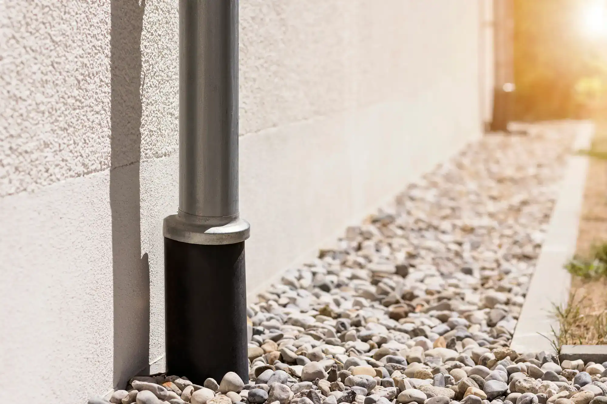 A metal downspout attached to a white building drains into a black splash block, surrounded by small gray and white pebbles—perfectly installed by an expert Excavation Contractor in Suffolk County, NY—with sunlight shining in the background.