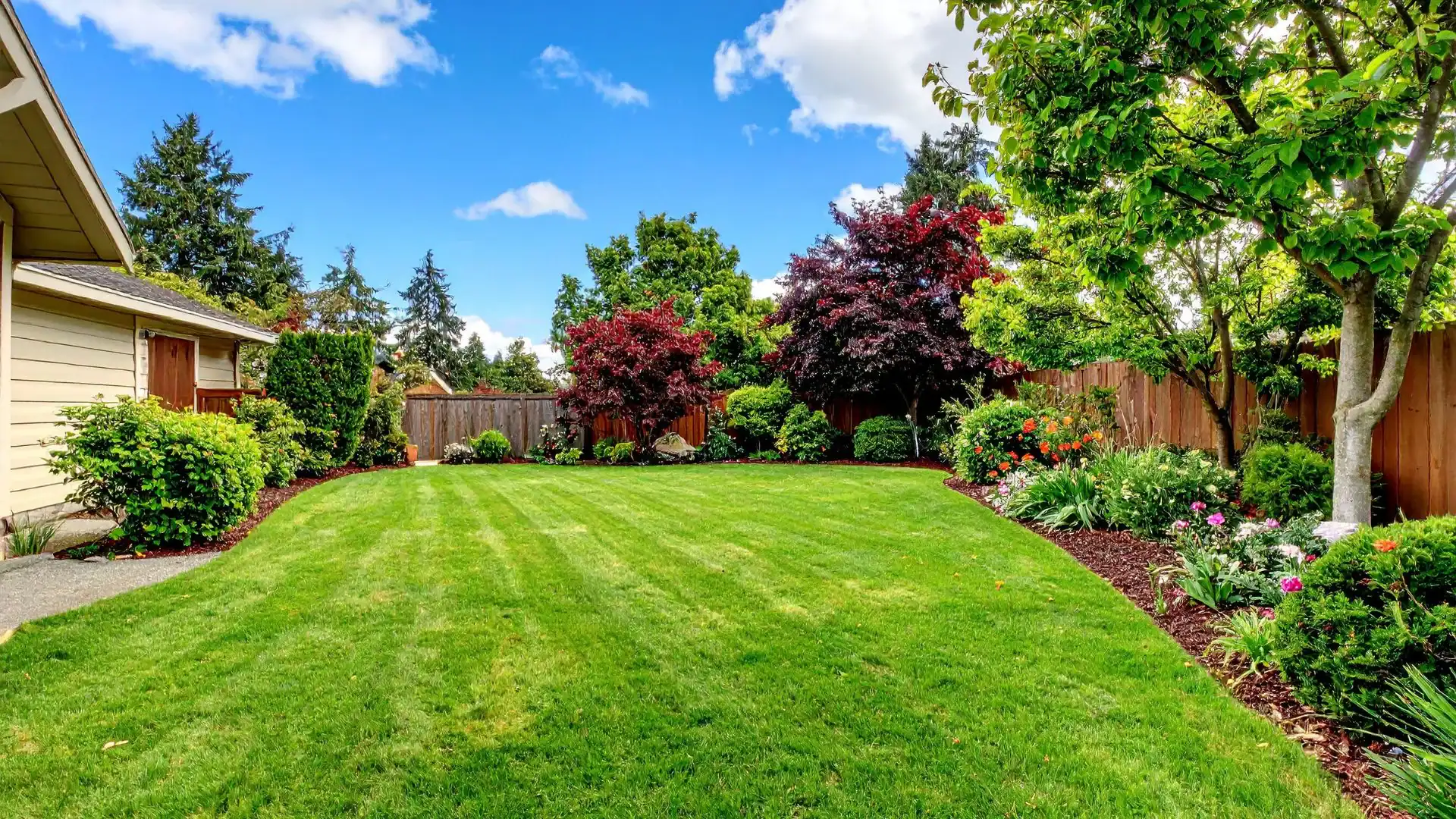 A backyard with a well-manicured green lawn, surrounded by trees, shrubs, and flower beds. A wooden fence lines the yard—perfect inspiration for any NY homeowner or anyone seeking an Excavation Contractor Suffolk County trusts.